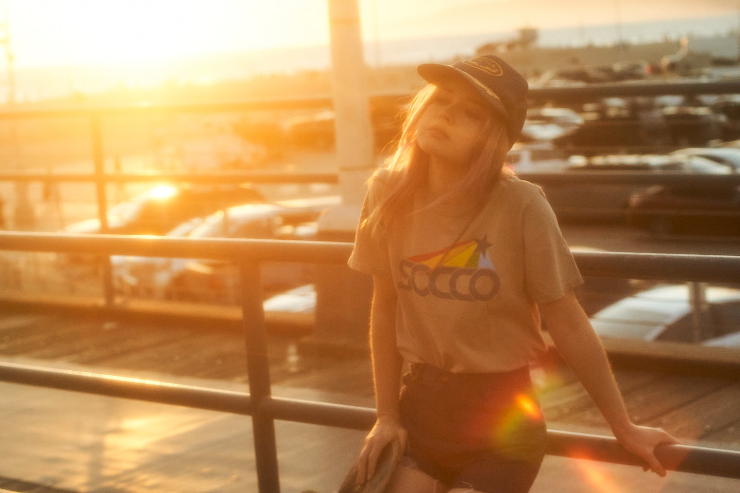 Female wearing a SOCCO t-shirt and shorts with a skateboard at sunset on a boardwalk in Los Angeles.