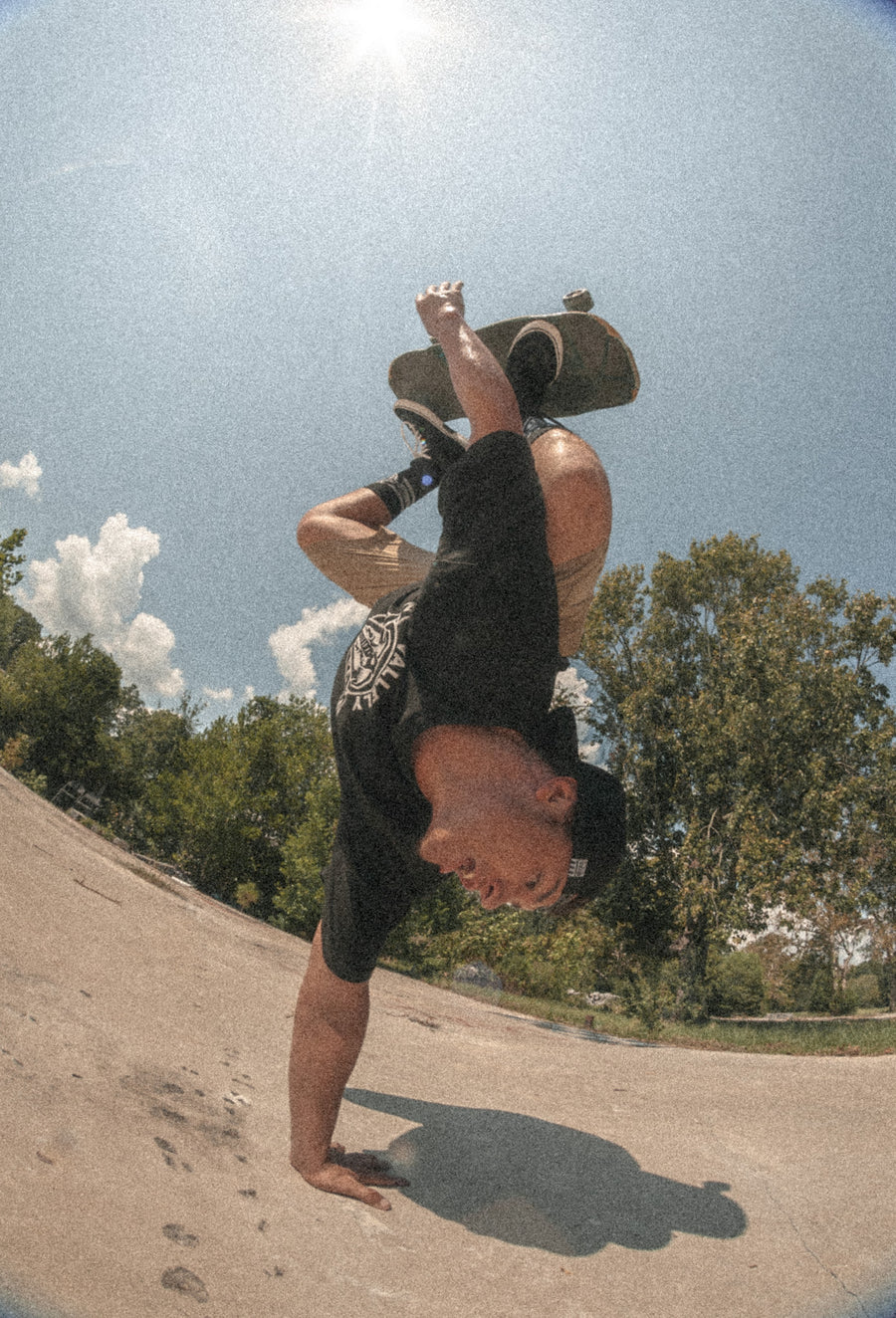 Skateboarder in shorts, black Dirty Donny x Mike Vallely Logo Tee, backwards Dirty Donny x Mike Vallely Cap, and khaki shorts doing a one-handed headstand with his skateboard in the air. He is wearing the black colorway of the Dirty Donny x Mike Vallely Collaboration Crew Socks.