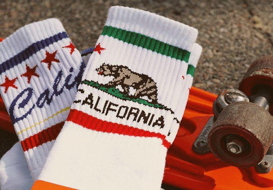 Two pairs of white athletic California themed SOCCO socks laid over an orange 1970s skateboard.