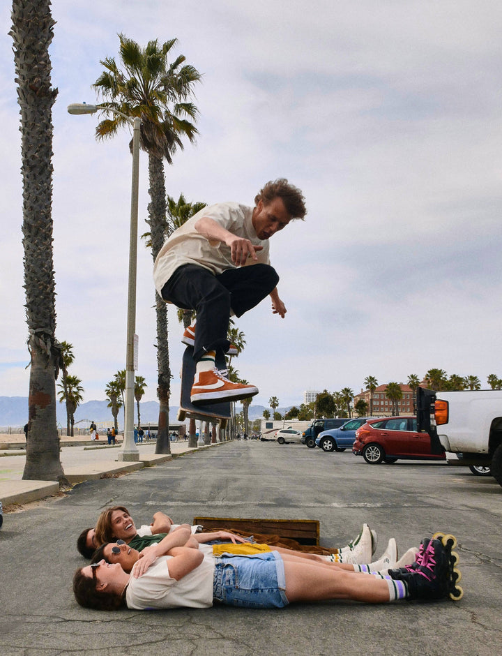 Male Skateboarder launching over friends laying on the asphalt.