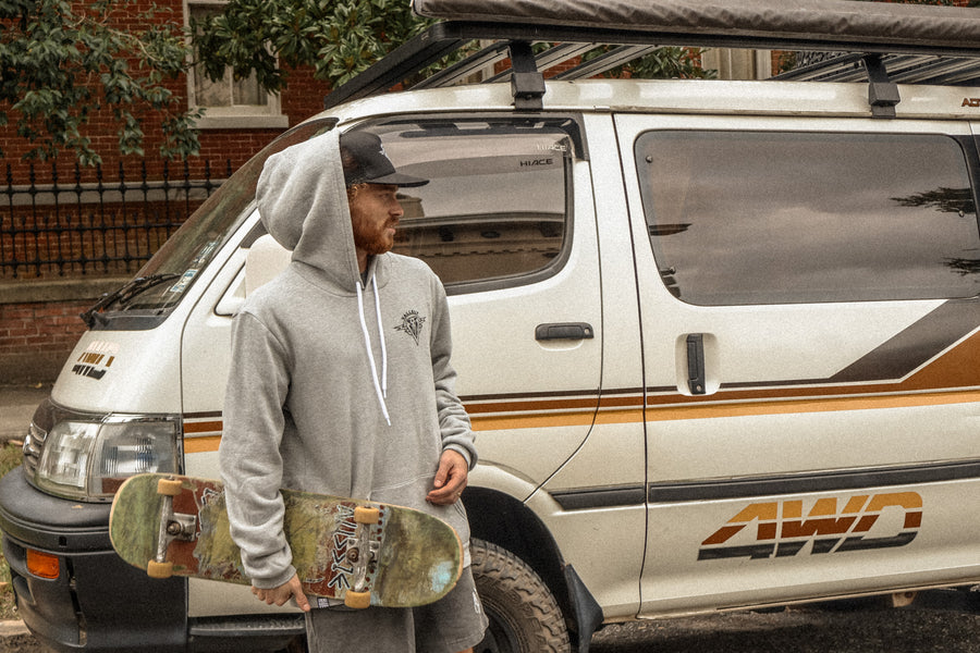 Skateboarder carrying his board in front of a retro striped van. He is wearing Dirty Donny x Mike Vallely Hoodie with the hood up and a Black Logo Cap.