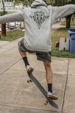 Skateboarder doing tricks on a sidewalk in the city while wearing the Dirty Donny x Mike Vallely Collaboration Heather Gray Hoodie and Black Logo Hat