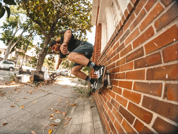 Skateboarder in Dity Donny x Mike Vallely Backwards cap, black logo tee, and black crew socks doing a wall ride on a brick wall.