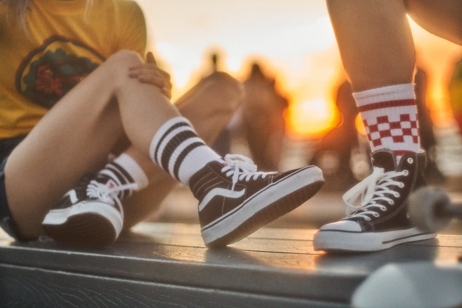 Female skateboarder wearing red checkered SOCCO socks with another female wearing SOCCO black striped white socks on a boardwalk at sunset.