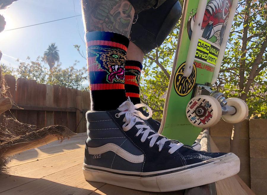 SOCCO x Dirty Donny Cougar Socks. Black Crew socks with three red stripes on the leg; on top of the stripes on the outside of the leg is a 70s style Cheetah head with it's mouth open, teeth bared, and tongue hanging out. The Cheetah is outlined in Acid Green. Dirty Donny Logo Signature on the top of the foot is in white.
